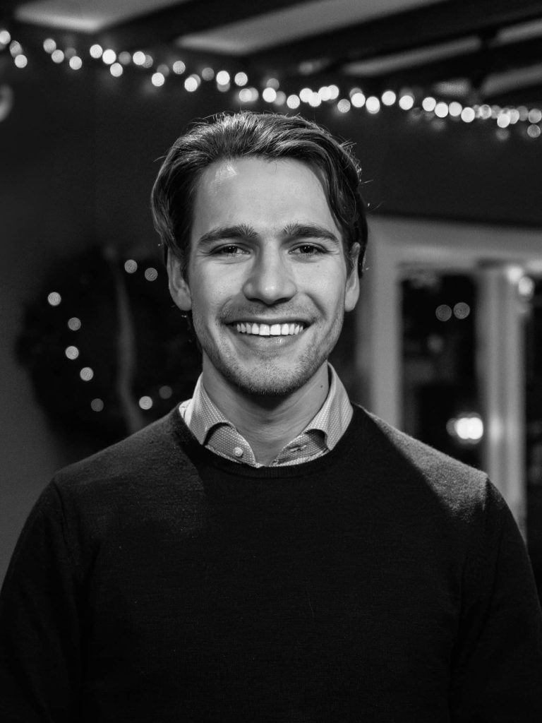 Cas Bierkens in een donkere polo met overhemd, staand in een open ruimte met sfeerverlichting op de achtergrond, kijkt met brede lach naar de camera.